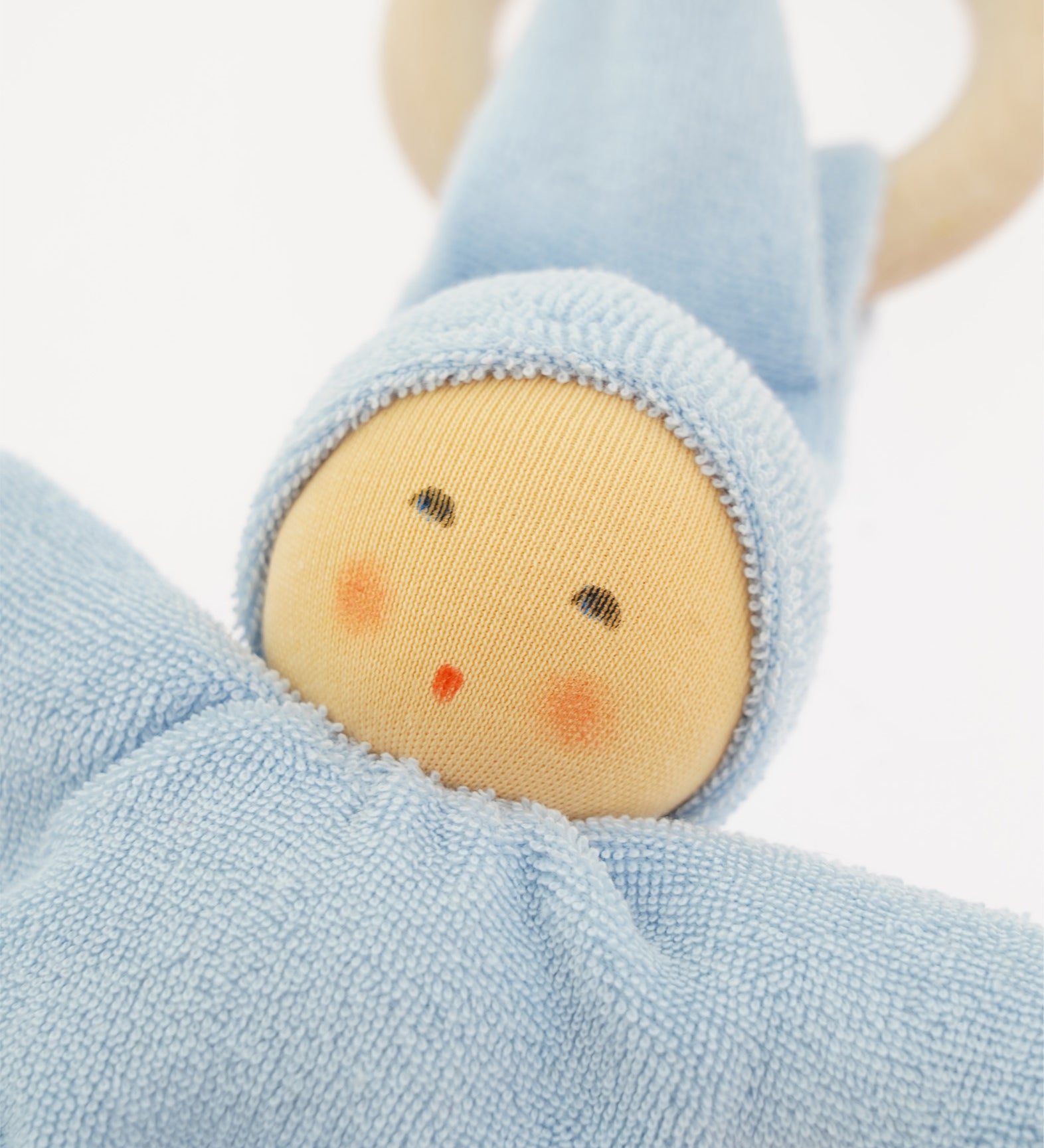 Close-up of the Nanchen Pimpel Doll Baby Comforter in soft blue. The doll's face features gently painted eyes, cheeks, and lips, framed by a light blue organic cotton terry hood. A wooden teething ring is partially visible behind the pointed hat.