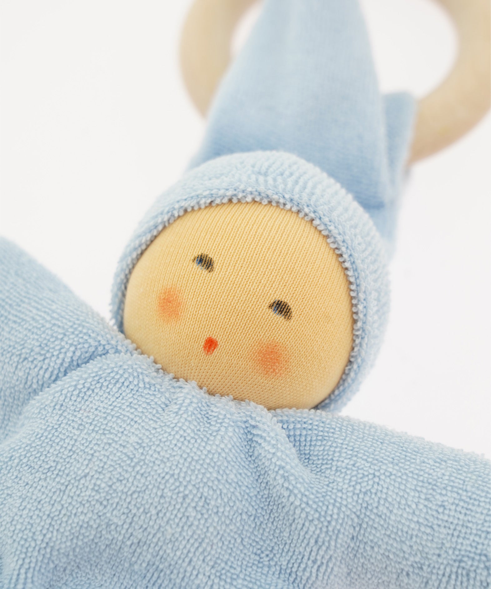 Close-up of the Nanchen Pimpel Doll Baby Comforter in soft blue. The doll's face features gently painted eyes, cheeks, and lips, framed by a light blue organic cotton terry hood. A wooden teething ring is partially visible behind the pointed hat.