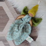 A Nanchen green Flower Fairy Ring Grasper placed on a rug on a wooden floor. A soft toy attached to a wooden ring that can be used as a grasping toy or hung up.