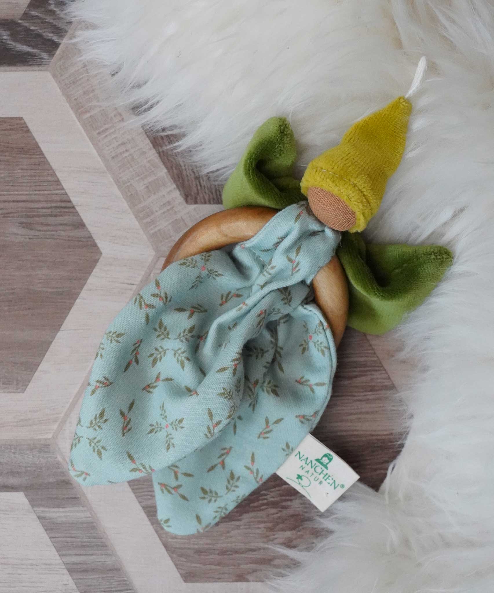 A Nanchen green Flower Fairy Ring Grasper placed on a rug on a wooden floor. A soft toy attached to a wooden ring that can be used as a grasping toy or hung up.