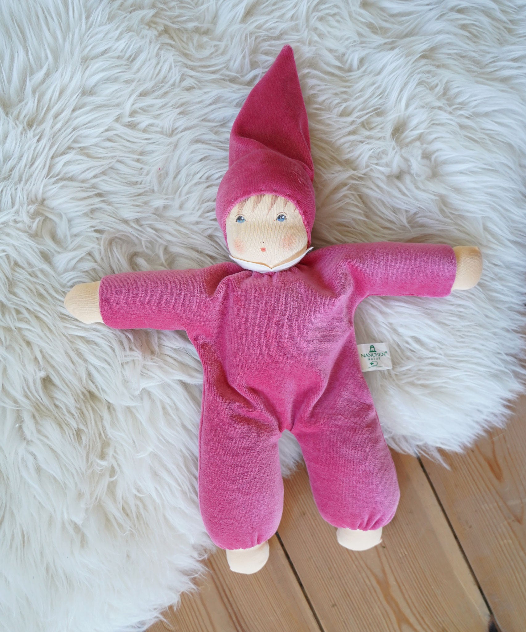 A pink Nanchen Nani Waldorf Doll placed on a sheepskin rug on a wooden floor.