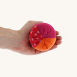 Nanchen Large Colourful, Organic Baby Rattle Ball on a hand for scale and colour. This is a soft terry material in red, orange and pink colours on a cream background.