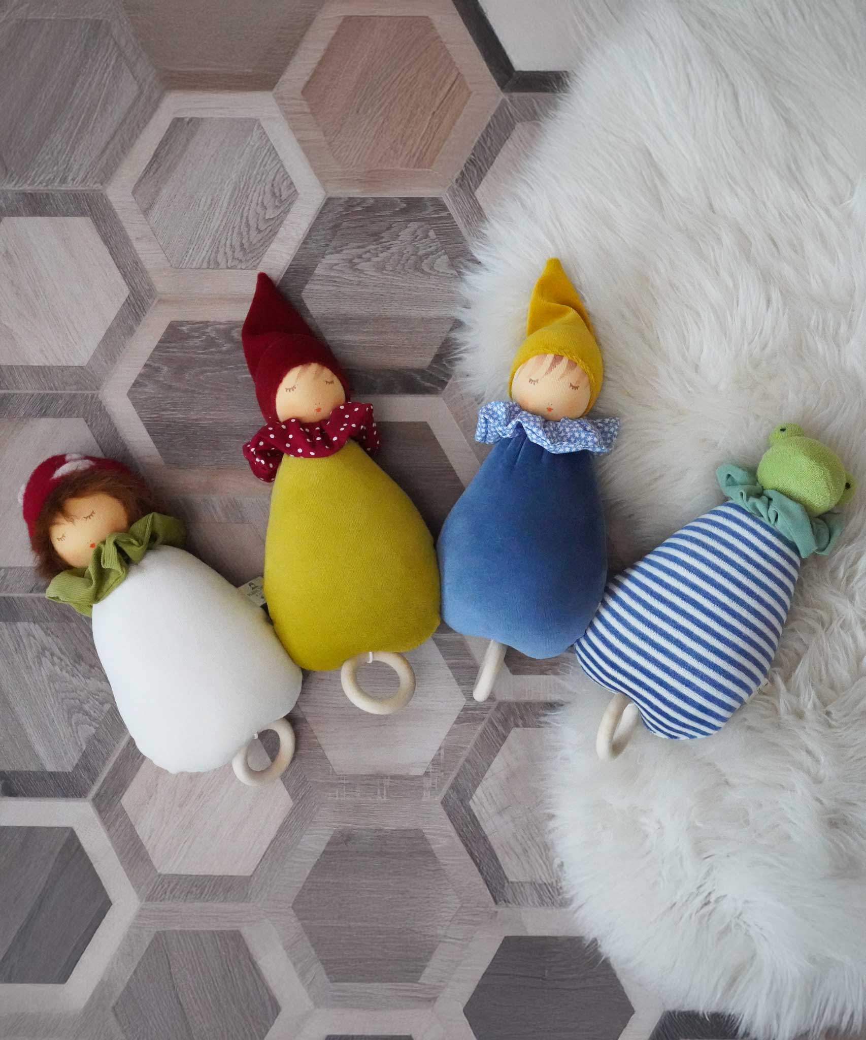 A collection of four Nanchen Lullaby Music Dolls. Musical Waldorf dollS, made from organic cotton and sheep's wool. The toadstool, little red riding hood, blue dream kings and frog lullaby dolls have been placed on a rug on a wooden floor. 