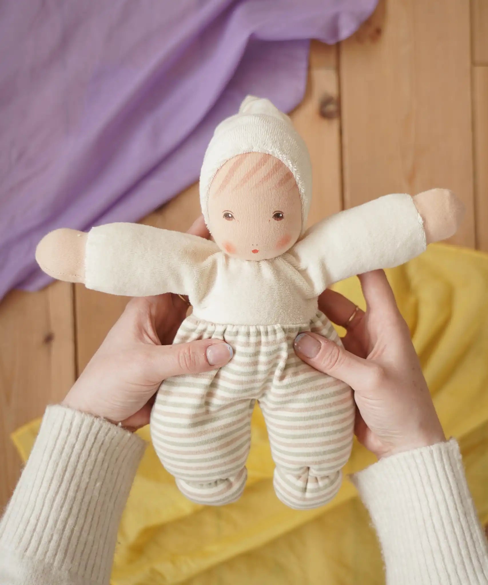 Nanchen Möpschen Waldorf doll in natural white colour with striped pattern on trousers in an adult's hand in the Babipur playroom.
