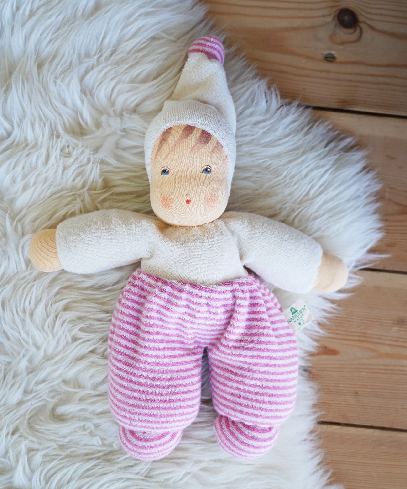 A Nanchen Möpschen Waldorf Doll with Pink Stripes placed on a sheepskin rug on a wooden floor.