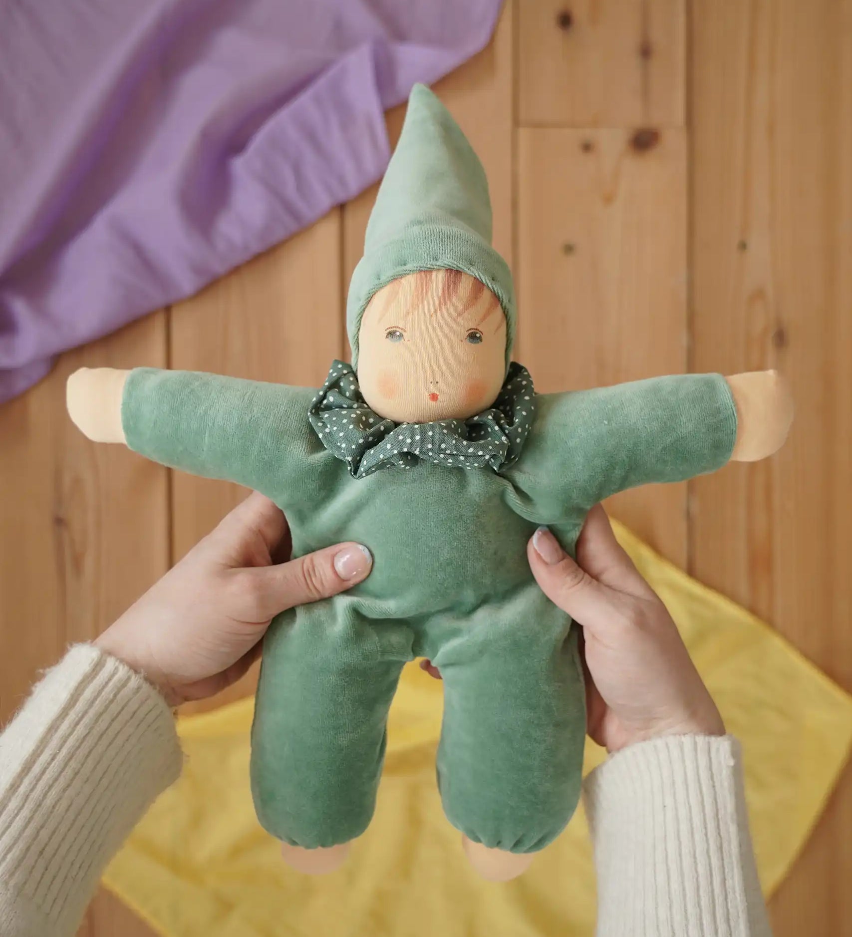 Nanchen Natur Waldorf Nani doll in mint green in an adult's hands in the Babipur playroom. Purple and yellow Laura's play cloths can be seen in the background.