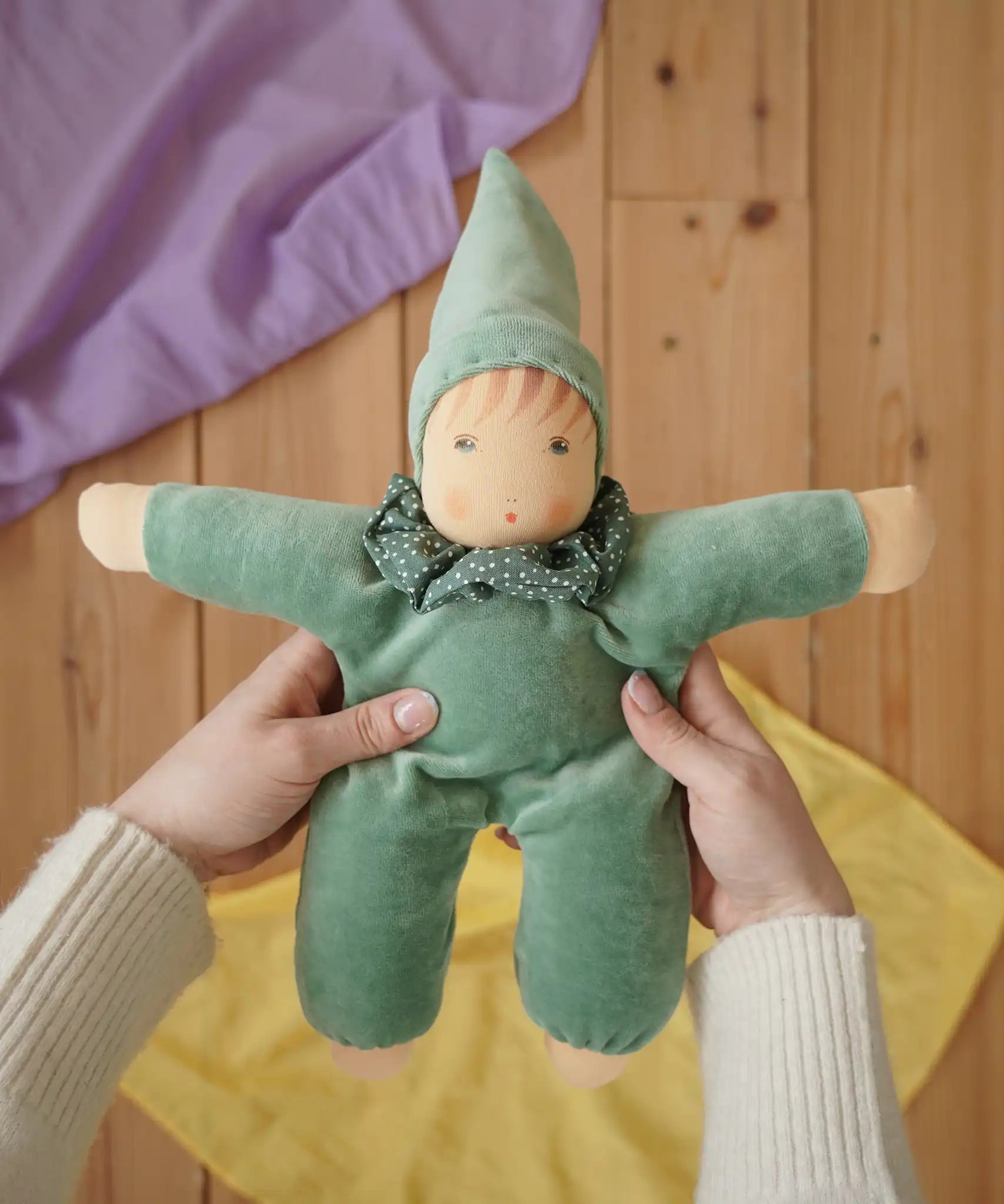Nanchen Natur Waldorf Nani doll in mint green in an adult's hands in the Babipur playroom. Purple and yellow Laura's play cloths can be seen in the background.
