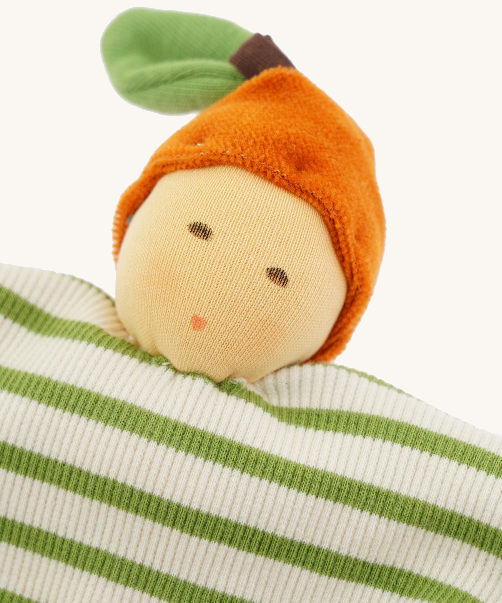 Close-up of the Nanchen Orange Baby Comforter, a handmade doll with a soft, peach-colored face, embroidered features, and an orange velour hat shaped like a fruit with a green leaf. The doll's body is made from organic cotton terry in green and white stripes.
