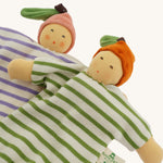 Two Nanchen baby comforters lying side by side: the Orange Baby Comforter in green and white stripes with an orange fruit hat, and the Peach Baby Comforter in lilac and white stripes with a pink fruit hat. Both dolls have soft, hand-embroidered faces and knotted feet.
