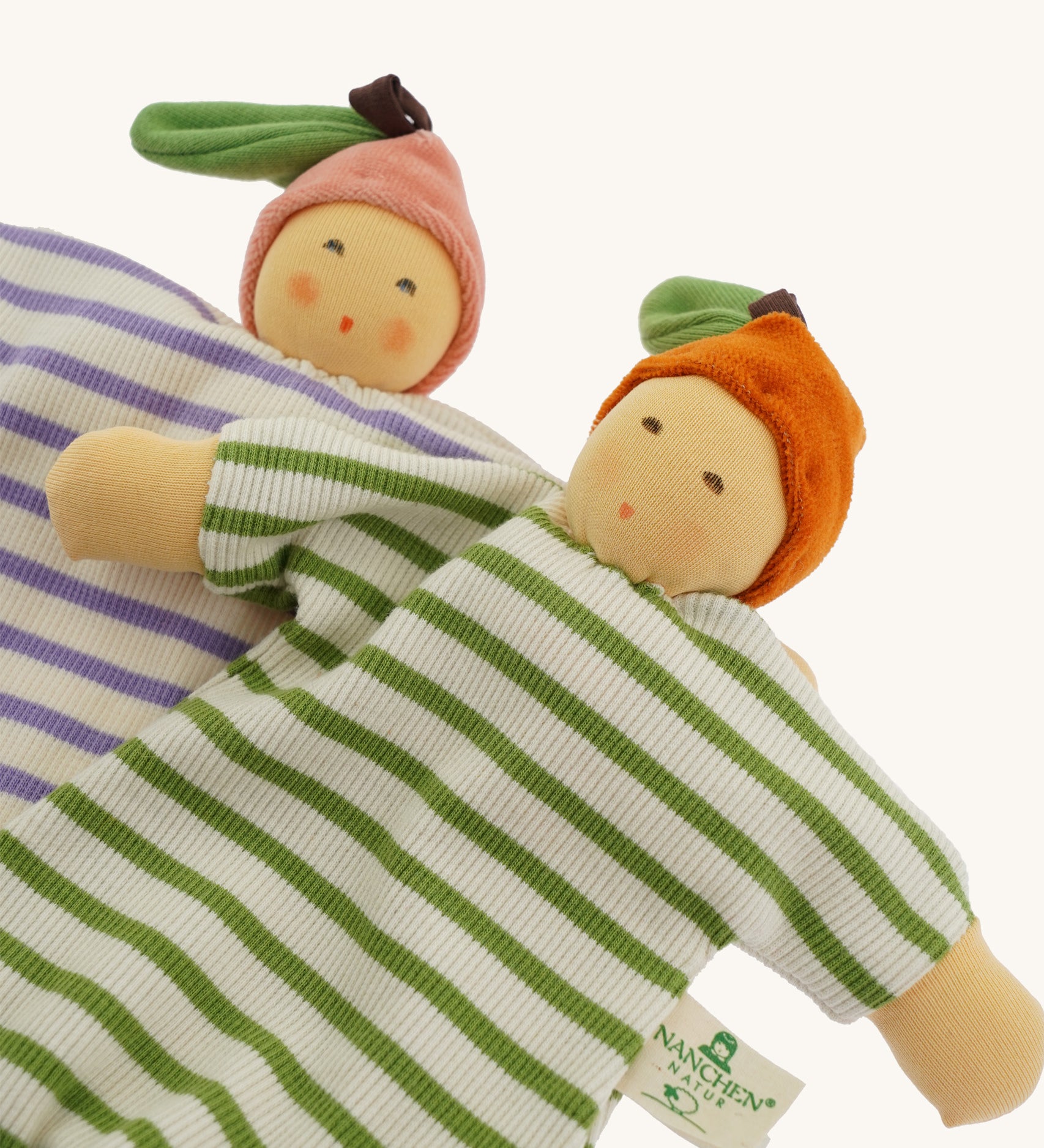 Two Nanchen baby comforters lying side by side: the Orange Baby Comforter in green and white stripes with an orange fruit hat, and the Peach Baby Comforter in lilac and white stripes with a pink fruit hat. Both dolls have soft, hand-embroidered faces and knotted feet.
