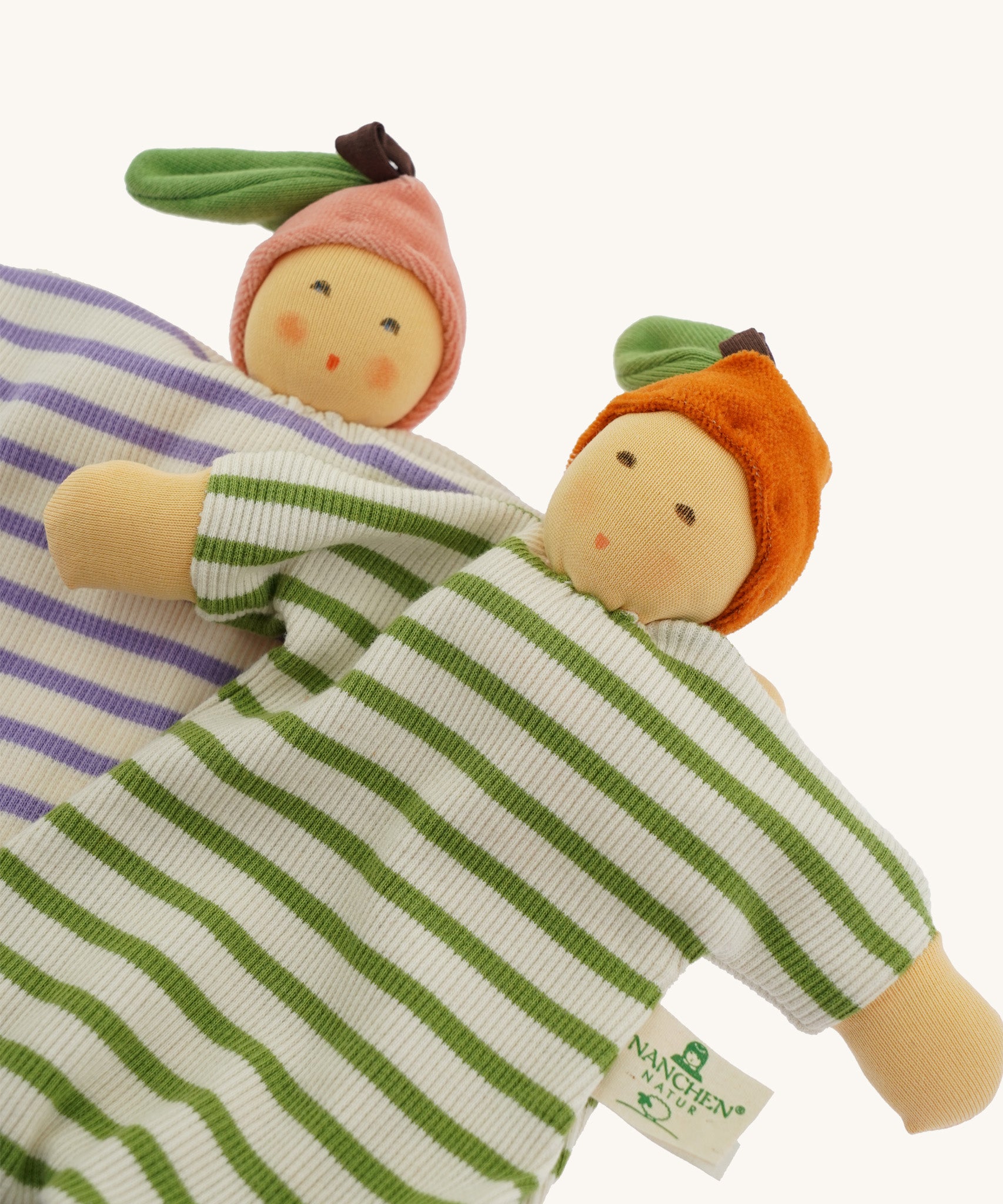 Two Nanchen baby comforters lying side by side: the Orange Baby Comforter in green and white stripes with an orange fruit hat, and the Peach Baby Comforter in lilac and white stripes with a pink fruit hat. Both dolls have soft, hand-embroidered faces and knotted feet.
