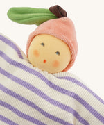 Close-up of the Nanchen Peach Baby Comforter, featuring a warm, peach-toned face with rosy cheeks and embroidered eyes and mouth. The doll wears a pink velour fruit-shaped hat with a green leaf and brown stem, and has a soft cotton body with lilac and white stripes.
