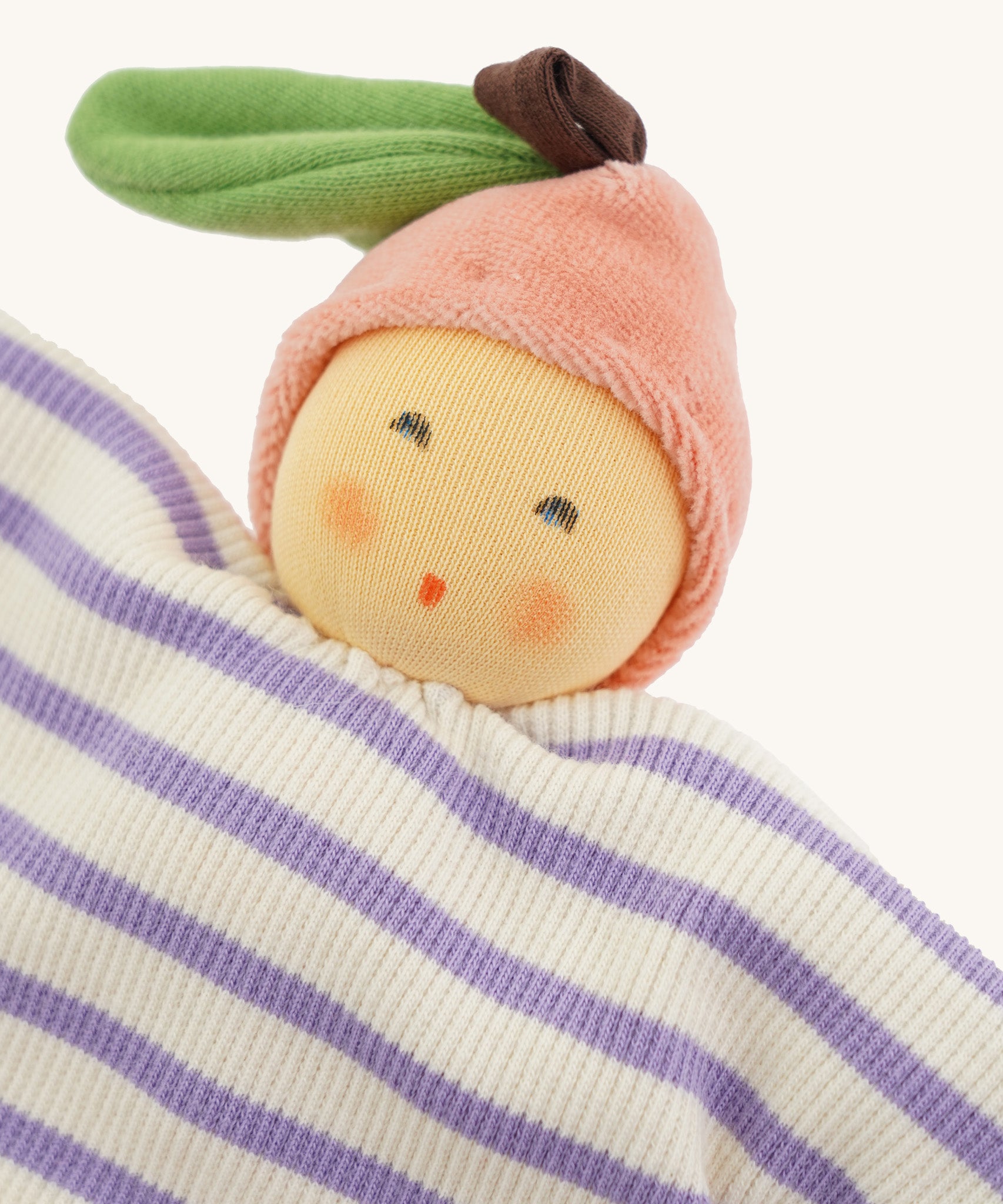Close-up of the Nanchen Peach Baby Comforter, featuring a warm, peach-toned face with rosy cheeks and embroidered eyes and mouth. The doll wears a pink velour fruit-shaped hat with a green leaf and brown stem, and has a soft cotton body with lilac and white stripes.
