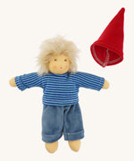 The Nanchen Frederik organic Waldorf doll dressed in a blue striped shirt and soft blue trousers, with the red gnome-style hat placed beside it. The doll’s blond mohair hair is fluffy and unstyled, and its hand-painted features are visible.
