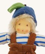 Close-up of the face of the Nanchen organic Waldorf doll Bela Blueberry. The doll has pale skin, hand-painted facial features, rosy cheeks, and wears a blue fruit-style hat with a green leaf and stem over messy blond hair.