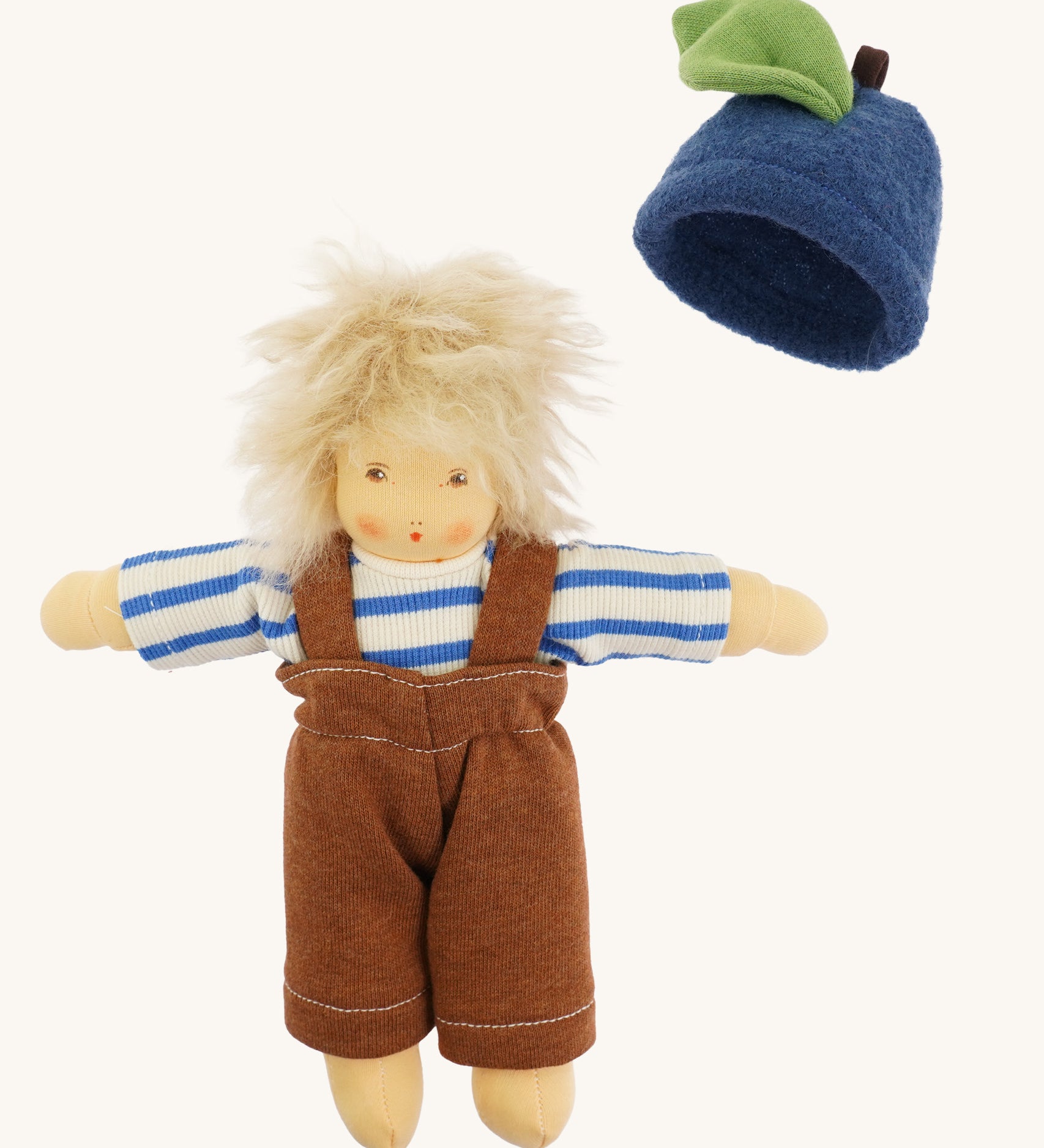Nanchen organic Waldorf doll dressed in the blue and white striped top and brown dungarees, with the blueberry fruit hat beside it. The doll has pale skin and fluffy platinum blond hair.