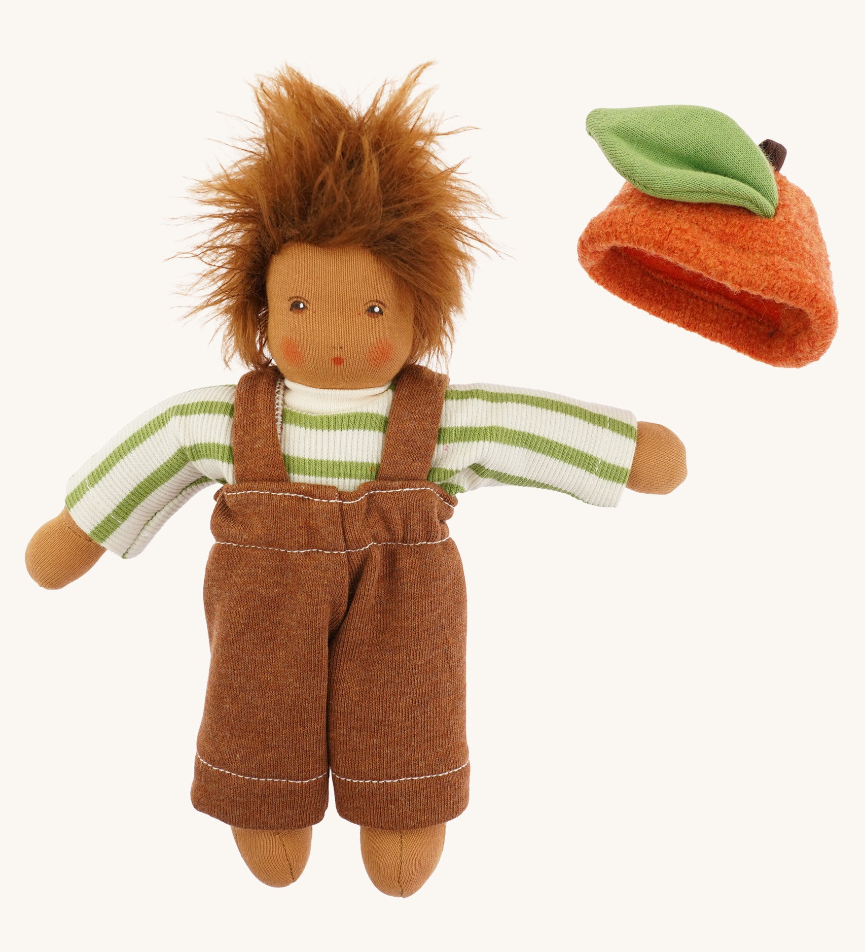 Nanchen organic Waldorf doll dressed in a green and white striped top and brown dungarees, with the orange fruit hat placed beside it. The doll has brown skin and fluffy brown hair.