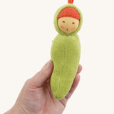 The Nanchen Pea Pod Rattle Doll is shown being held in an adult’s hand, demonstrating its small, baby-friendly size. The soft green terry body and hand-painted face are visible, along with the red top knot detail.