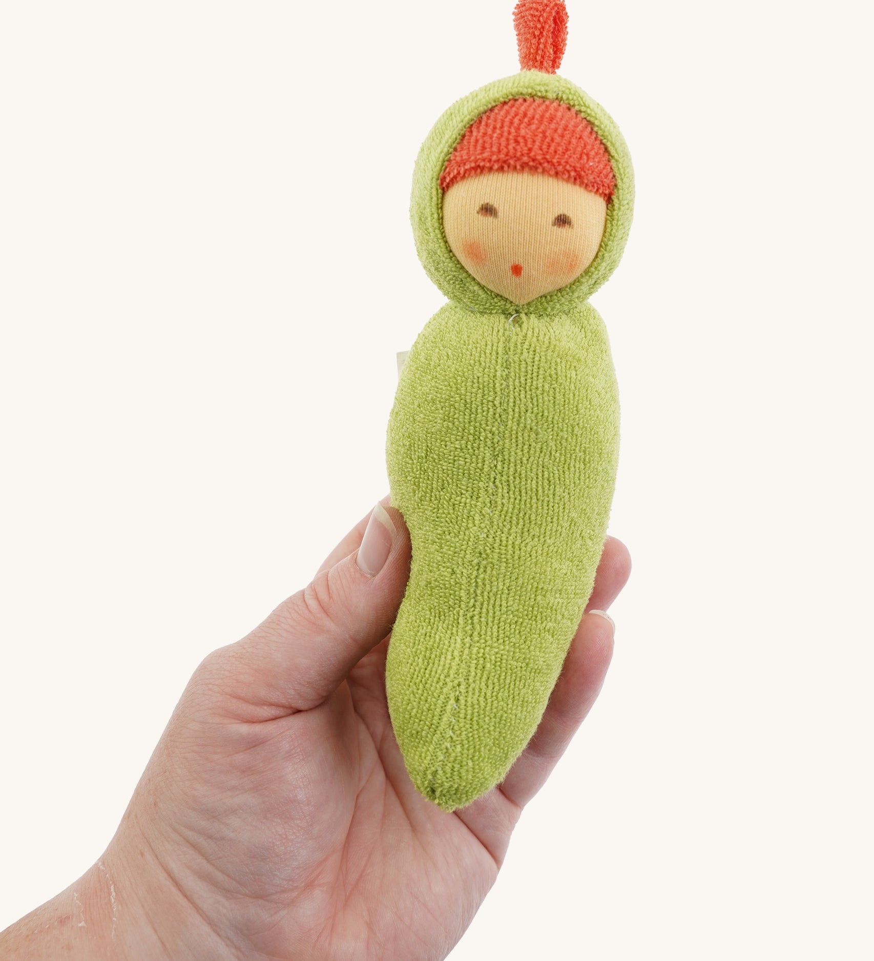 The Nanchen Pea Pod Rattle Doll is shown being held in an adult’s hand, demonstrating its small, baby-friendly size. The soft green terry body and hand-painted face are visible, along with the red top knot detail.