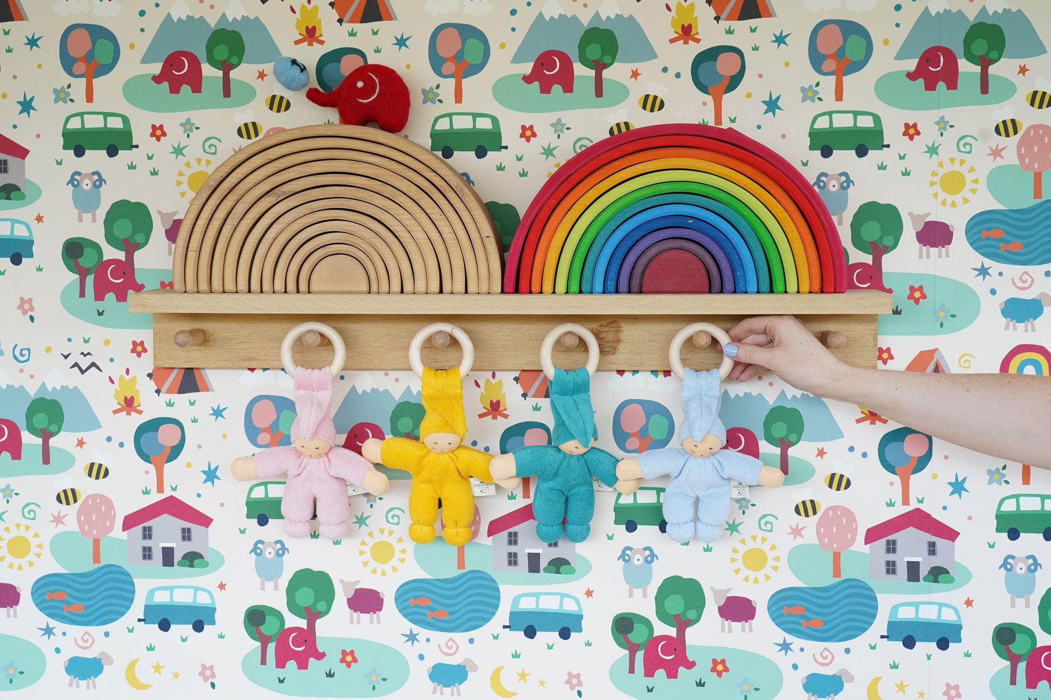 Four Pimpel doll comforters hung up by their wooden hops on wooden pegs on a shelf. There are pink, yellow, turquoise and light blue dolls. 