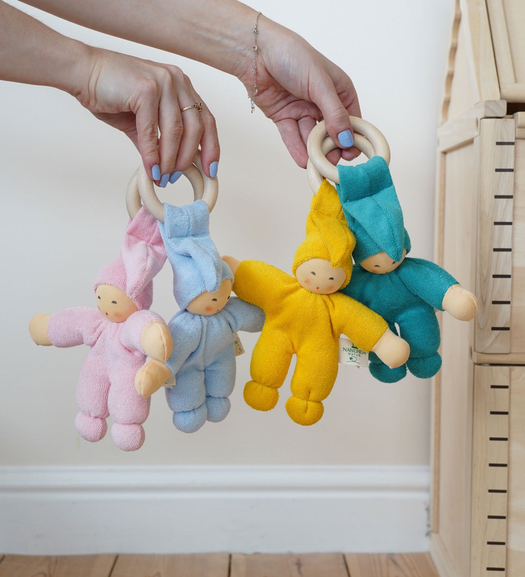 Four Pimpel doll baby comfortmers being held up in an adult's hands. There is a pink, light blue, yellow and turquoise doll. 