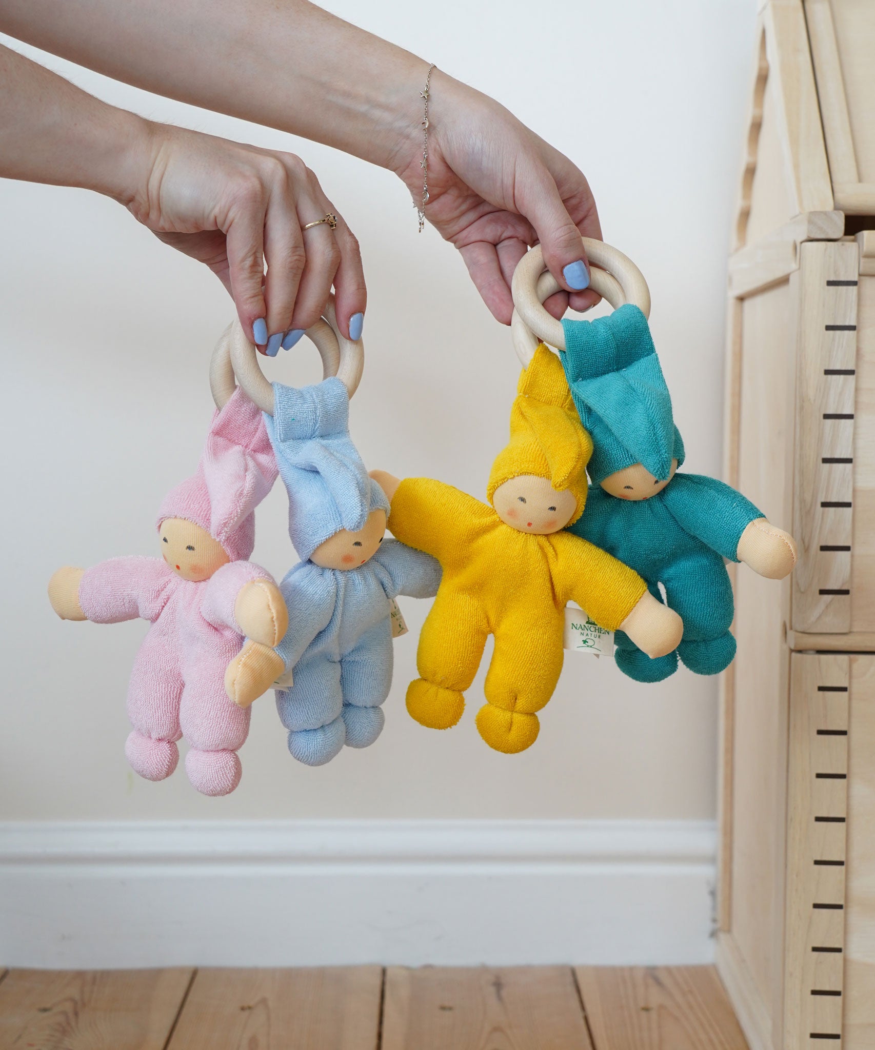 Four Pimpel doll baby comfortmers being held up in an adult's hands. There is a pink, light blue, yellow and turquoise doll. 