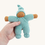The Nanchen Pimpel Waldorf Doll in sage green is shown being held in an adult’s hand, highlighting its small, baby-friendly size. The doll’s face is hand-painted and it has a soft terry cloth body with a gentle expression.
