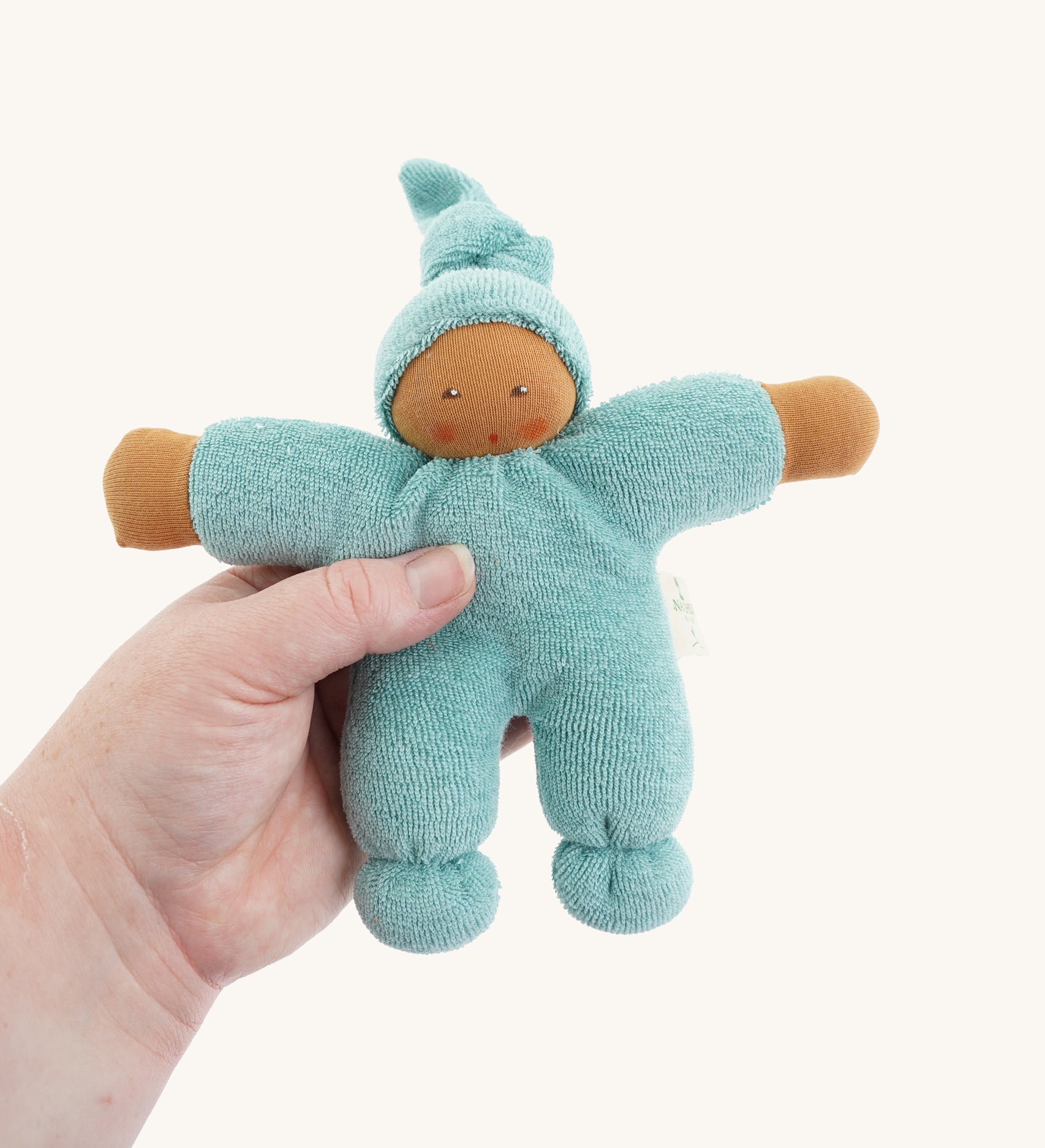 The Nanchen Pimpel Waldorf Doll in sage green is shown being held in an adult’s hand, highlighting its small, baby-friendly size. The doll’s face is hand-painted and it has a soft terry cloth body with a gentle expression.