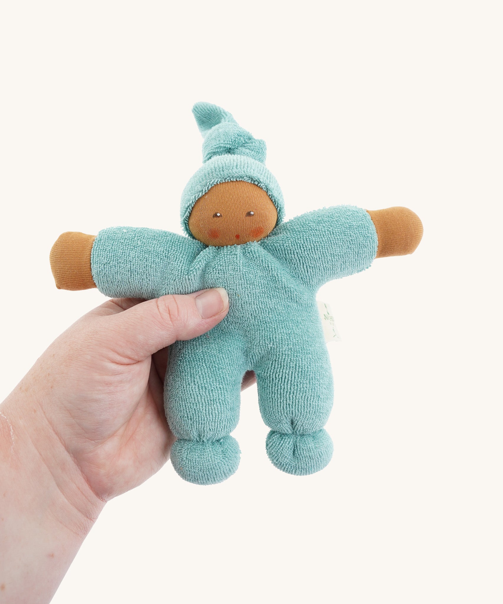 The Nanchen Pimpel Waldorf Doll in sage green is shown being held in an adult’s hand, highlighting its small, baby-friendly size. The doll’s face is hand-painted and it has a soft terry cloth body with a gentle expression.