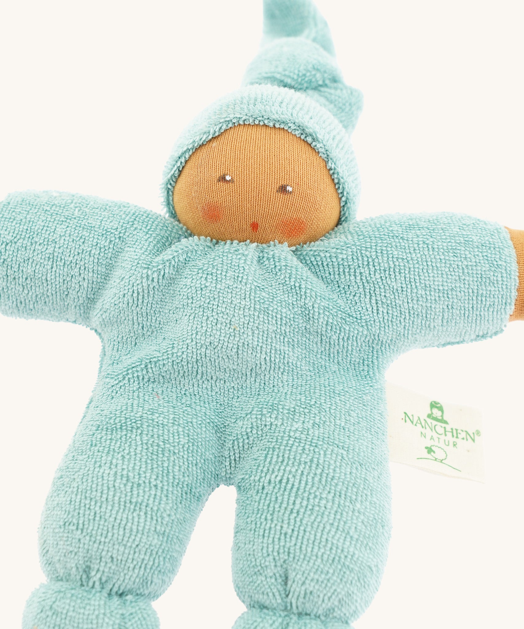 Close-up of the Nanchen Pimpel Waldorf Doll in sage green, showing its soft terry cotton body and sweet hand-painted facial features. The doll wears a pointed hood and has rosy cheeks, with a "Nanchen Natur" tag on its side.