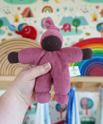 A Nanchen Pimpel Waldorf Handcrafted Doll with a berry pink coloured outfit and matching knotted hat being held up in an adult's hand in the Babipur playroom.