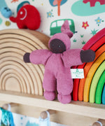 A Nanchen Pimpel Waldorf Handcrafted Doll with a berry pink coloured outfit an matching knotted hat. The doll has been placed on a wooden shelf in the Babipur playroom in front of two Grimm's wooden rainbow stacking toys, one is natural and one is rainbow coloured. 