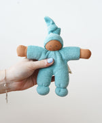 The Nanchen Pimpel Waldorf Doll in sage green is shown being held in an adult’s hand, highlighting its small, baby-friendly size. The doll’s face is hand-painted and it has a soft terry cloth body with a gentle expression.
