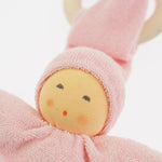 Close-up of the Nanchen Pimpel Doll in pastel pink. The doll’s sweet painted face shows soft blush and dot eyes, framed by a pink terry cloth hood. A wooden teething ring is looped through the pointed hat just behind the head.