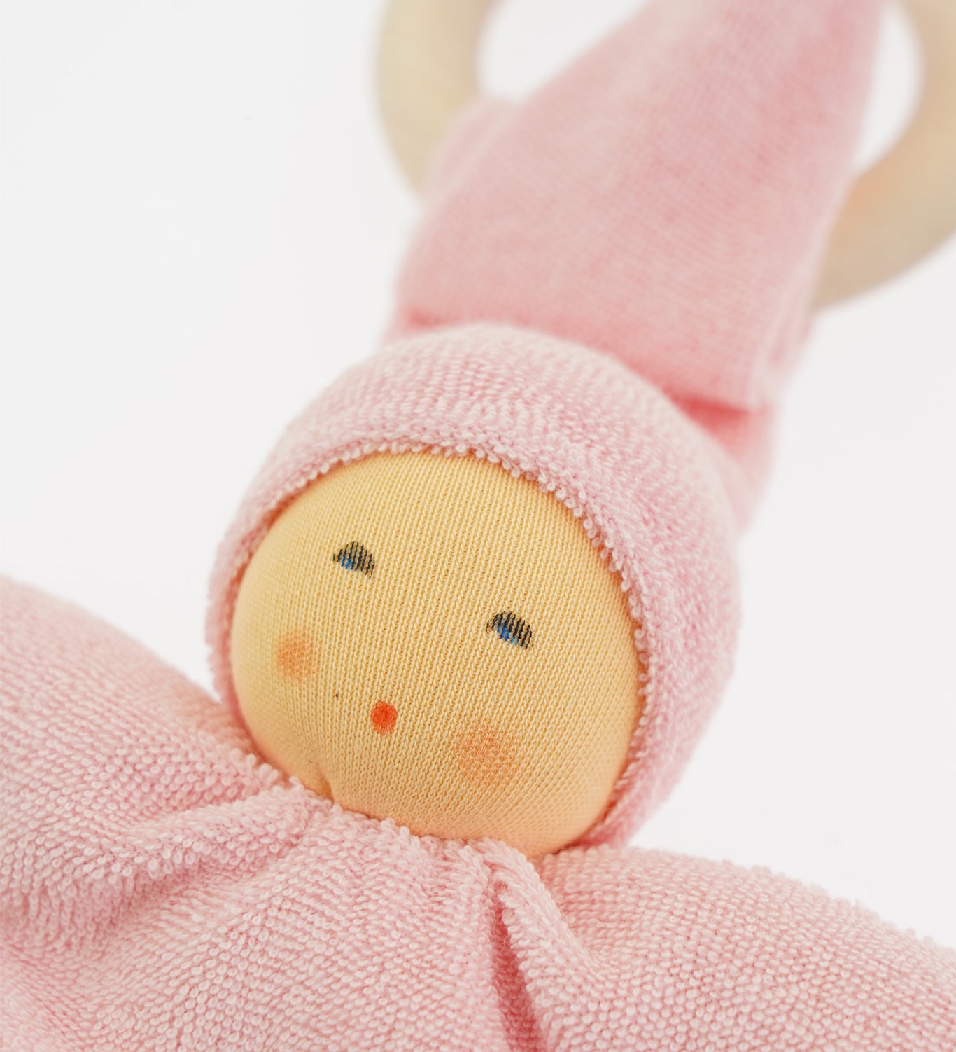 Close-up of the Nanchen Pimpel Doll in pastel pink. The doll’s sweet painted face shows soft blush and dot eyes, framed by a pink terry cloth hood. A wooden teething ring is looped through the pointed hat just behind the head.