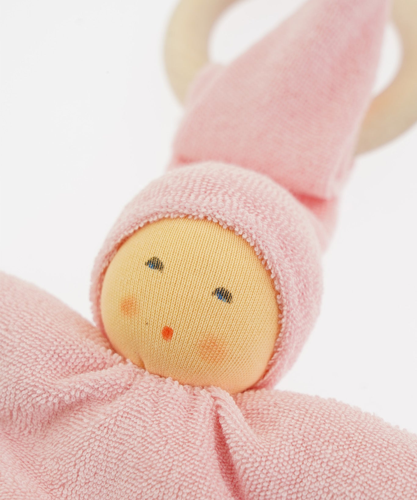 Close-up of the Nanchen Pimpel Doll in pastel pink. The doll’s sweet painted face shows soft blush and dot eyes, framed by a pink terry cloth hood. A wooden teething ring is looped through the pointed hat just behind the head.