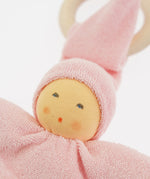 Close-up of the Nanchen Pimpel Doll in pastel pink. The doll’s sweet painted face shows soft blush and dot eyes, framed by a pink terry cloth hood. A wooden teething ring is looped through the pointed hat just behind the head.