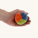 Nanchen small Colourful, Organic Baby Rattle Ball on a hand for scale and showing the different colours. This is a soft terry material in blue, orange and green colours on a cream background.