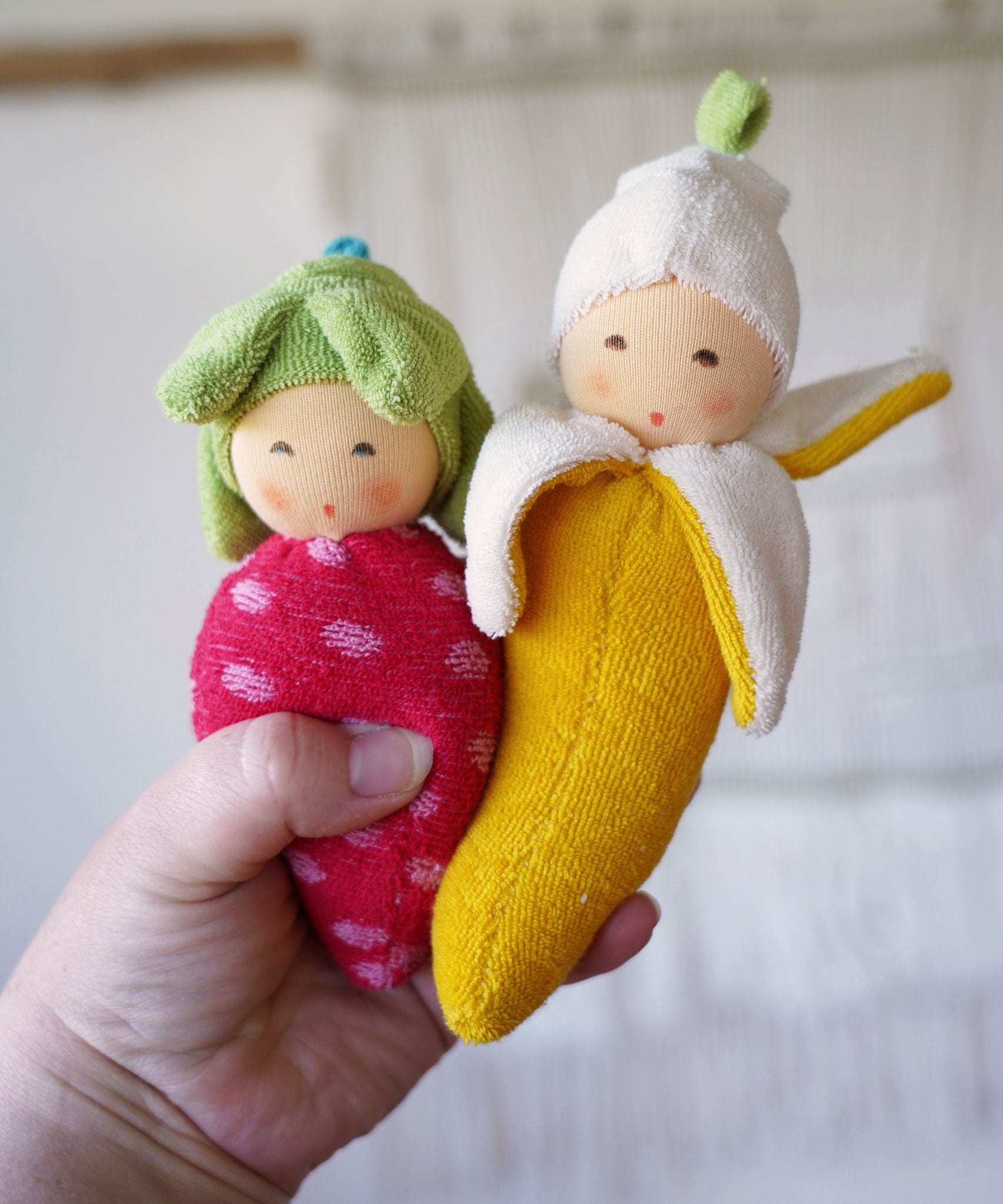 Nanchen banana and strawberry Baby Rattle Dolls in an adult's hand. 