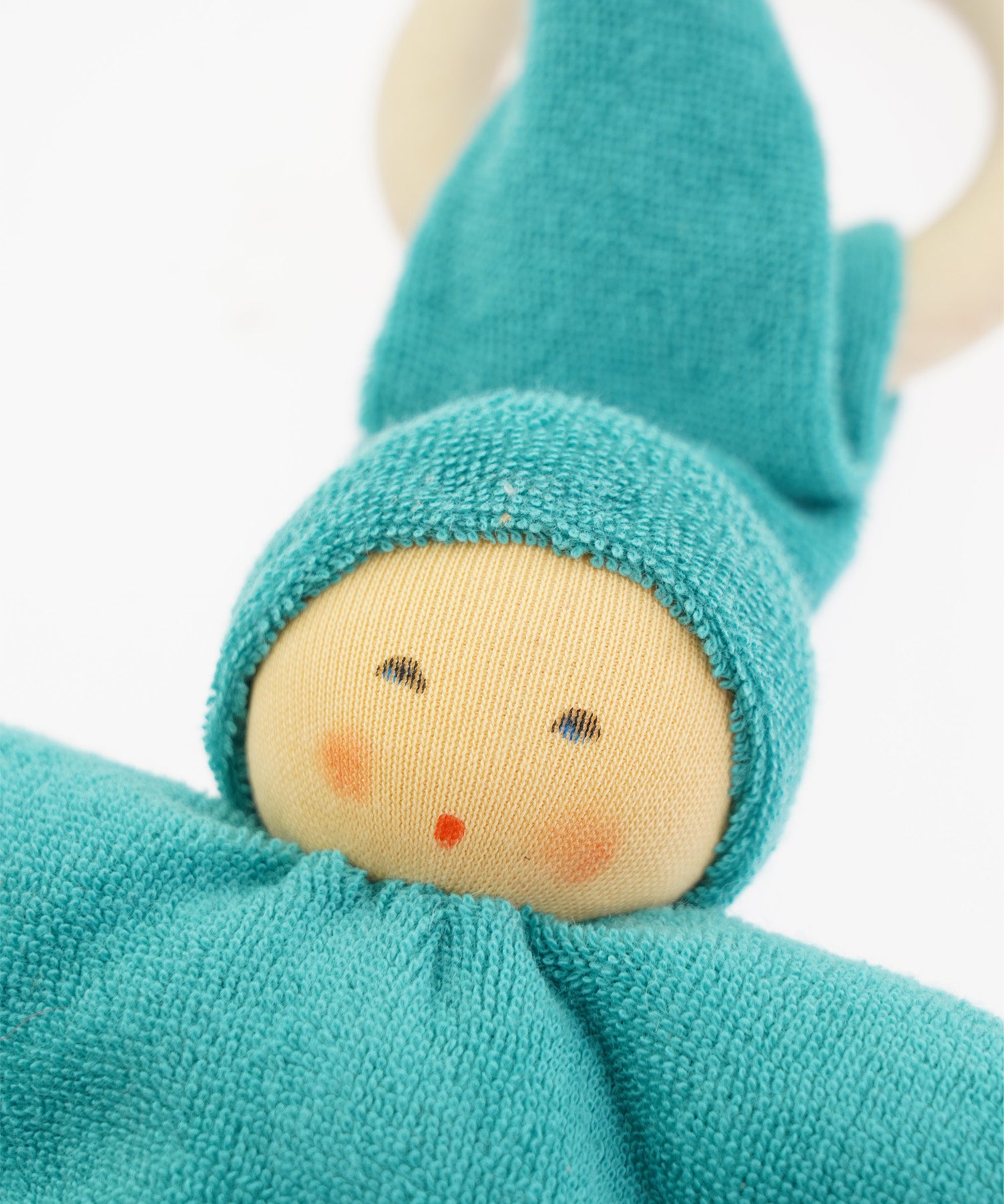 Close-up of the Nanchen Pimpel Doll in vibrant turquoise. The doll's painted facial features include soft blush cheeks and a red mouth, peeking out from under a cozy turquoise hood made of organic cotton terry. A wooden teething ring is looped through the pointed hat.
