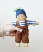 A Organic Handcrafted Nanchen Blueberry Child Waldorf Doll called Bella. The doll has a removable outfit which consists of a blue and white striped top, brown dungaree style bottoms and a blueberry hat.