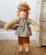 A Nanchen Flore Waldorf Doll being held up in an adult's hand. The Plan Toys Victorian dolls house can be seen just off to the side. 