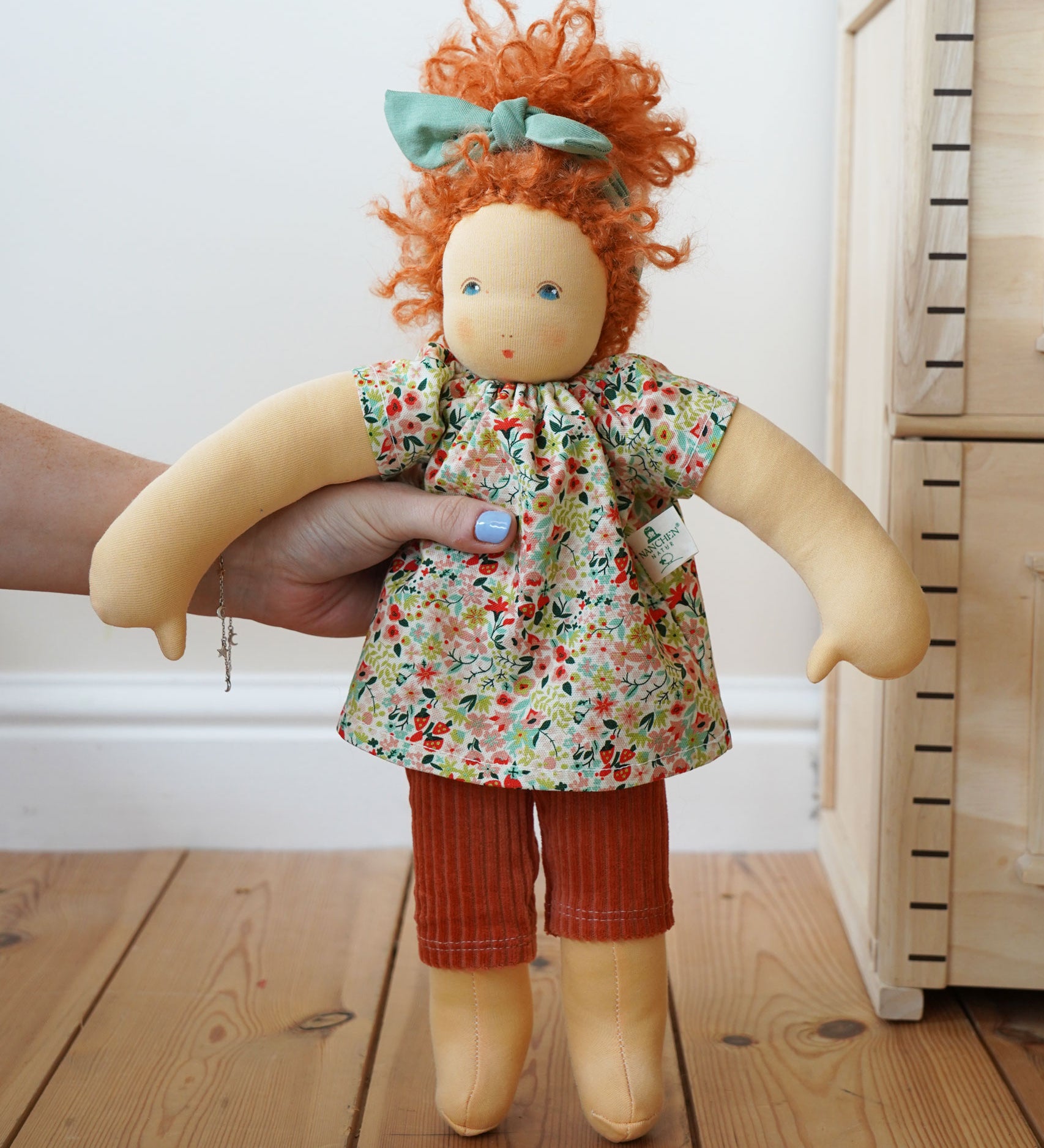 A Nanchen Flore Waldorf Doll being held up in an adult's hand. The Plan Toys Victorian dolls house can be seen just off to the side. 