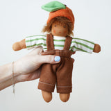 A Nanchen Otto Orange Child Waldorf Doll held up in an adult's hand. The doll is wearing e green and white striped top with brown coloured dungaree style bottoms and a orange hat The dolls outfit can be removed.