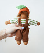 A Nanchen Otto Orange Child Waldorf Doll held up in an adult's hand. The doll is wearing e green and white striped top with brown coloured dungaree style bottoms and a orange hat The dolls outfit can be removed.