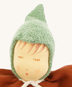 Close-up of the Nanchen Walnut Baby Doll's face, showing delicate hand-painted details including closed eyes with lashes, subtle blush, and soft lips. The doll wears a textured green hood made of organic cotton terry.