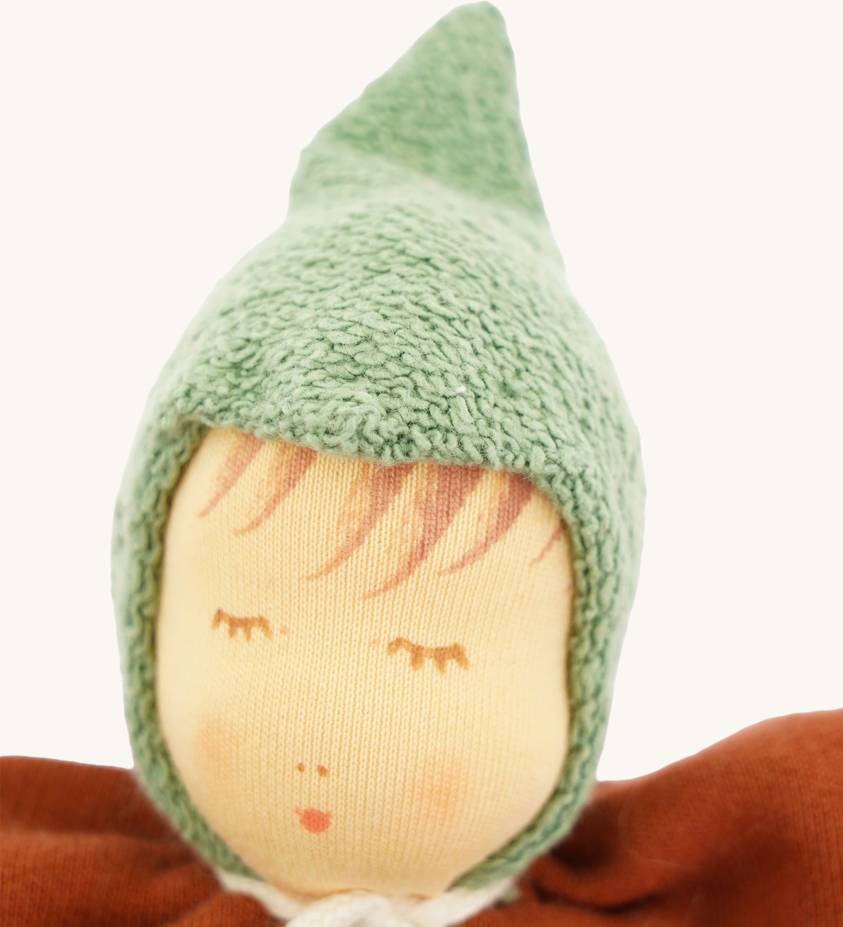 Close-up of the Nanchen Walnut Baby Doll's face, showing delicate hand-painted details including closed eyes with lashes, subtle blush, and soft lips. The doll wears a textured green hood made of organic cotton terry.