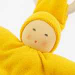 Close-up of the yellow Nanchen Waldorf Pimpel Doll. The doll has a hand-painted face with rosy cheeks and a small red mouth, surrounded by a bright yellow cotton terry hood and body. A natural wooden teething ring is seen in the background.