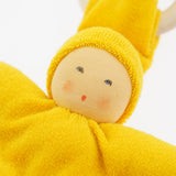 Close-up of the yellow Nanchen Waldorf Pimpel Doll. The doll has a hand-painted face with rosy cheeks and a small red mouth, surrounded by a bright yellow cotton terry hood and body. A natural wooden teething ring is seen in the background.