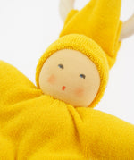 Close-up of the yellow Nanchen Waldorf Pimpel Doll. The doll has a hand-painted face with rosy cheeks and a small red mouth, surrounded by a bright yellow cotton terry hood and body. A natural wooden teething ring is seen in the background.